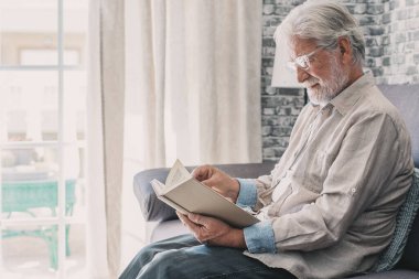 Headshot portrait close up of old happy and relaxed man sitting reading a book at home. Mature male person enjoying free time having fun indoor.