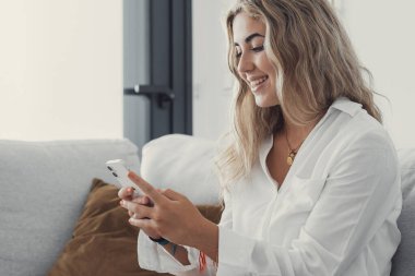 Çekici, sarışın bir kadının portresi. Kanepede cep telefonu kullanıyor. Evde internette geziniyor. Çevrimiçi konsept yaşam tarzı rahatlıyor