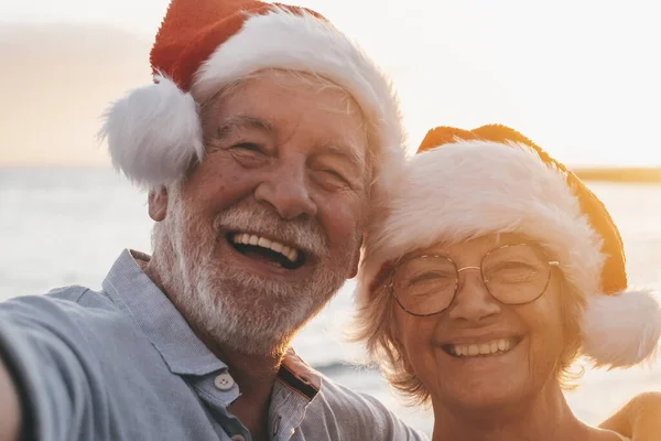 Two old happy seniors wearing christmas hats at the beach taking a selfie of them smiling and having fun with the sunset at the background at evening. Cute couple of old persons looking at the camera enjoying vacations.