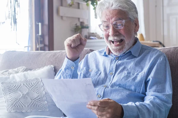 Excited elderly man celebrate health insurance deal closing looking and reading the medical results. Happy mature senior feel overjoyed make sign good bank agreement with specialist. Checking outcomes at home.