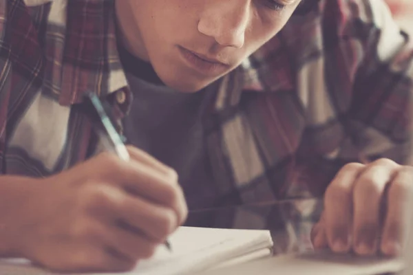 caucasian teenager indoor doing homework on the table at home - blonde guy writing and reading in his laptop or computer to get greats scores - portrait of boy with headphones