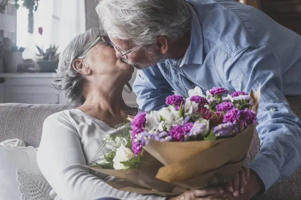 Old man giving flowers at his wife sitting on the sofa at home for the San Valentines day. Pensioners enjoying surprise kissing together. In love people having fun.