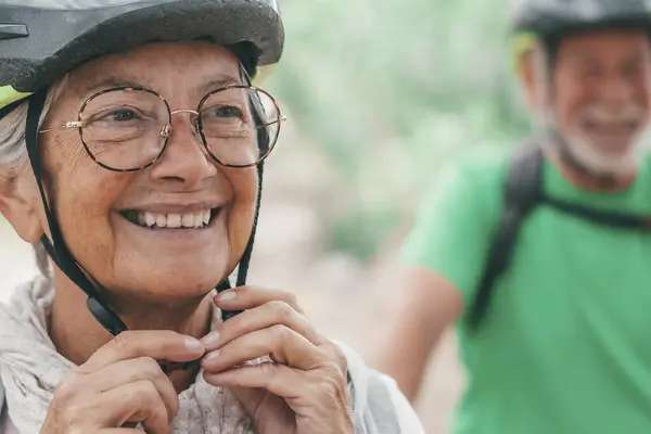 Portrait of one old woman smiling and enjoying nature outdoors riding bike with her husband laughing. Headshot of mature female with glasses feeling healthy. Senior putting on helmet to go trip with bicycles.