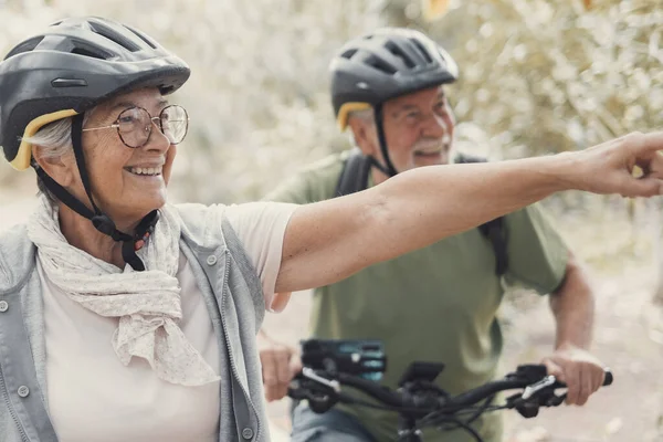 Two happy old mature people enjoying and riding bikes together to be fit and healthy outdoors. Active seniors having fun training in nature. Portrait of one old man smiling in a bike trip with his wife. Woman indicating something and looking at it.