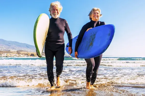 two old and mature people having fun and enjoying their vacations outdoors at the beach wearing wetsuits and holding a surfboard to go surfing in the water with waves - active senior smiling and doing water sports