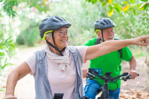 Two happy old mature people enjoying and riding bikes together to be fit and healthy outdoors. Active seniors having fun training in nature. Portrait of one old man smiling in a bike trip with his wife. Woman indicating something and looking at it.