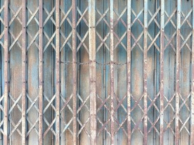 Katlanabilir metal kapı yaşlanmış ve yıpranmış, paslı yüzeyine kazınmış sessiz hikayelerle nöbet tutuyor, dayanıklılığa işaret ediyor.