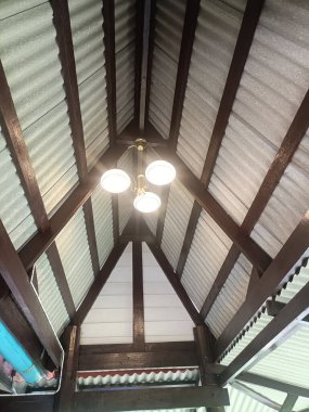 Görüntü, merkezden sarkan, ahşap kirişler ve eğri büğrü metal çatı panelleriyle çevrili, avize lamba armatürüne sahip yüksek tavanı gösteriyor.