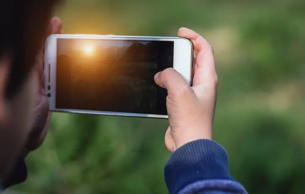 Little boy or a girl holding a smartphone using both hands and taking a photo of sunset. Colorful sunny outdoors background, travel concept, close up.
