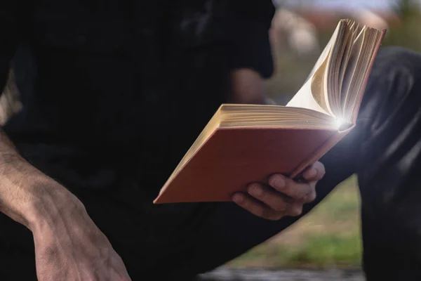 Man sitting and reading a book outdoor. Male hand holding a book outside. Student studying in nature or priest reading a bible. Focus on the book, copy space, close up.