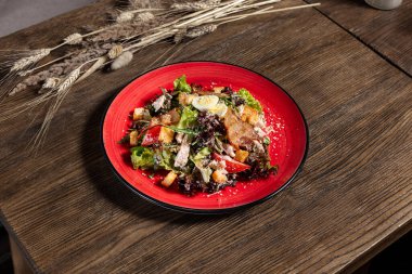 Koyu ahşap arka planda kırmızı seramik tabakta kızarmış domuz pastırması ve kızarmış tavuklu Sezar salatası..