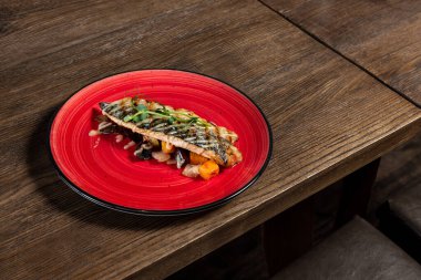 Hardal soslu ızgara uskumru filetosu ve fırında pişirilmiş sebzelerin üzerine mikroyeşillikler serpiştirilmiş. Yiyecek kırmızı seramik bir tabakta, koyu, ahşap bir arka planda yatıyor..