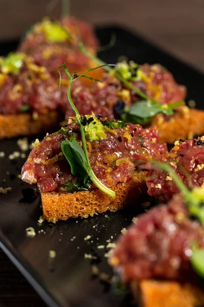 Beef tartare garnished with microgreens and a slice of avocado, topped with toasted buckwheat bread sauteed in garlic butter. Food lies on a rectangular ceramic plate on a wooden background.