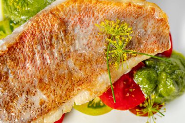 Yeşil ve kırmızı domatesli ızgara levrek fileto ve pesto sosu. Yiyecek hafif bir zemin üzerinde seramik bir tabakta yatıyor..