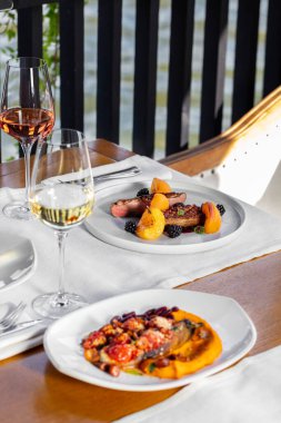 Beyaz şarapla üç çeşit yemek. Domatesli patlıcan, sebzeli levrek ve böğürtlen soslu ördek bifteği ahşap bir masada. Yakınlarda bir bardak beyaz şarap var..