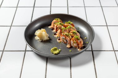 Somonlu suşi, karides tempura ve avokado. Üstü unagi soslu ve Japon biberli mayonezli. Küçük yeşil soğan halkalarıyla süslenmiş. Suşi, hafif arka planda metal bir plakada yatar..