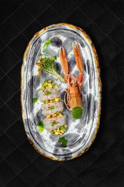 Langoustine Sashimi avokado tartartare 'nin üstüne acı biber dilimleri ve üstüne de soğan. Yakınlarda deniz kabuğu ve mikroyeşil var. Yiyecek seramik bir tabakta, siyah bir zemin üzerinde duruyor..