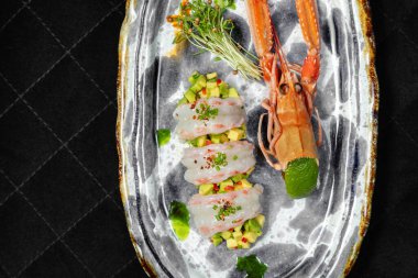 Langoustine Sashimi avokado tartartare 'nin üstüne acı biber dilimleri ve üstüne de soğan. Yakınlarda deniz kabuğu ve mikroyeşil var. Yiyecek seramik bir tabakta, siyah bir zemin üzerinde duruyor..