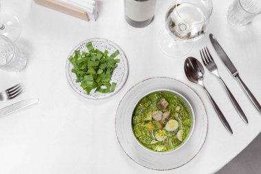 Dana eti, patates, ıspanak ve bıldırcın yumurtalı yeşil borş çorbası. Çorba hafif bir seramik tabağa dökülüyor, yanında yeşil bir kapak, bir şişe ve bir bardak beyaz şarap var..