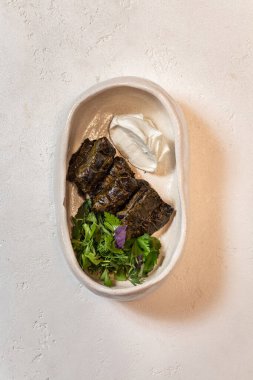 Dolma kuzu filetosu ve üzüm yapraklarına sarılmış bitkilerle pilav. Yakınlarda farklı yeşilliklerden ekşi krema ve karışık salata bulunur. Yemekler oval seramik tabakta yatıyor..