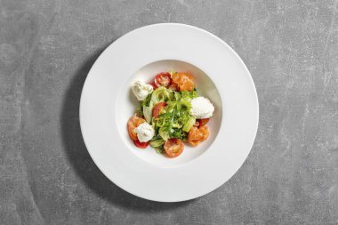 Arugula soslu yaz salatası, mozzarella, vişneli domates, soslu somon. Gri taşlı arka planda hafif bir seramik tabakta yiyecek yatıyor..