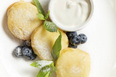 Yaban mersinli süzme peynir krepleri, nane yaprakları ve seramik tabakta pudra şekeri. Yakınlarda ekşi kremalı sos kasesi var. Tabak gri bir taş yüzeyde yatıyor..