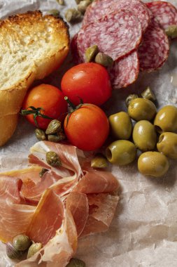 İtalyan aperatifleri prosciutto, salam, zeytin, baget tostları ve kiraz domatesleri ahşap bir tahtanın üzerinde açık parşömen üzerinde. Tahta gri bir arka planda duruyor..