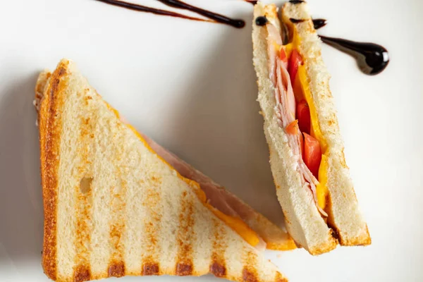 stock image Toasted bread sandwich with cheese, ham and tomatoes, cut in two and topped with balsamic sauce. The sandwich lies on a light ceramic plate on a gray stone background.