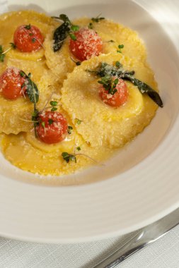 Mozzarella soslu Ravioli ve üstünde rendelenmiş parmesan olan vişneli domates. Yiyecek hafif bir seramik tabakta açık bir masa örtüsünde, yakınlarda birkaç bardak beyaz şarap ve bir vazo çiçek var..