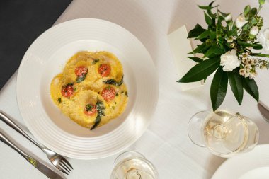 Mozzarella soslu Ravioli ve üstünde rendelenmiş parmesan olan vişneli domates. Yiyecek hafif bir seramik tabakta açık bir masa örtüsünde, yakınlarda birkaç bardak beyaz şarap ve bir vazo çiçek var..