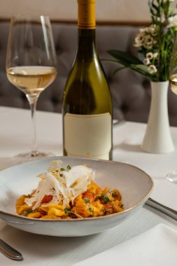 Domates soslu sebzeli Gnocchi. Üstünde mikroyeşiller ve karnabahar dilimleri var. Yiyecek hafif bir masa örtüsünün üzerindeki seramik bir tabakta yatıyor. Yakınlarda beyaz şarap bardakları ve bir vazo çiçek var..