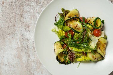 Sebze salatası, kabak, kiraz domatesleri, çam fıstığı, zeytinyağı ve balzamik sos. Yiyecek, renkli bir zemindeki açık seramik bir tabakta yatıyor..