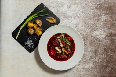 Ukrayna çorbası çorbası, hafif seramik tabakta mantar ve sebzelerle birlikte. Yakınlarda kızarmış patates ve yeşil soğanlı bir levha var. Tabaklar hafif kumaş arkaplanda durur.