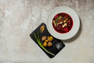 Ukrayna çorbası çorbası, hafif seramik tabakta mantar ve sebzelerle birlikte. Yakınlarda kızarmış patates ve yeşil soğanlı bir levha var. Tabaklar hafif kumaş arkaplanda durur.