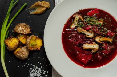 Ukrayna çorbası çorbası, hafif seramik tabakta mantar ve sebzelerle birlikte. Yakınlarda kızarmış patates ve yeşil soğanlı bir levha var. Tabaklar hafif kumaş arkaplanda durur.