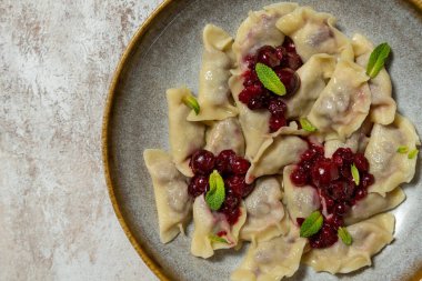 Kiraz ve böğürtlenli Ukrayna hamur tatlısı. Frenk üzümü, kiraz sosu ve nane yaprakları üstte. Yiyecek, açık renk bir arka planda, koyu bir seramik tabakta yatıyor..