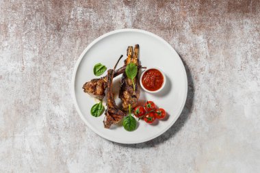 Üç parça ızgara kuzu pirzolası seramik tabakta duruyor. Yakınlarda vişneli domates, marul ve soslu sos var. Plaka hafif bir kumaş zemin üzerinde duruyor..