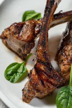 Üç parça ızgara kuzu pirzolası seramik tabakta duruyor. Yakınlarda vişneli domates, marul ve soslu sos var. Plaka hafif bir kumaş zemin üzerinde duruyor..