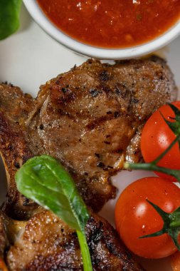 Üç parça ızgara kuzu pirzolası seramik tabakta duruyor. Yakınlarda vişneli domates, marul ve soslu sos var. Plaka hafif bir kumaş zemin üzerinde duruyor..