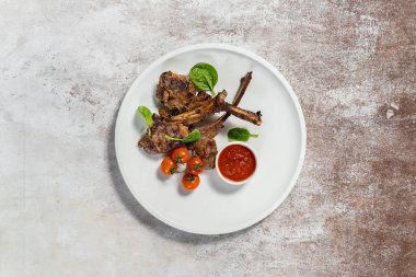 Üç parça ızgara kuzu pirzolası seramik tabakta duruyor. Yakınlarda vişneli domates, marul ve soslu sos var. Plaka hafif bir kumaş zemin üzerinde duruyor..