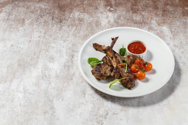 Üç parça ızgara kuzu pirzolası seramik tabakta duruyor. Yakınlarda vişneli domates, marul ve soslu sos var. Plaka hafif bir kumaş zemin üzerinde duruyor..