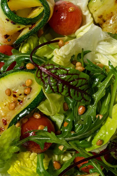 Vegetable mix salad with zucchini, cherry tomatoes, pine nuts, olive oil and balsamic sauce. Food lies in a light ceramic plate on a colored background.