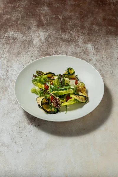 Vegetable mix salad with zucchini, cherry tomatoes, pine nuts, olive oil and balsamic sauce. Food lies in a light ceramic plate on a colored background.