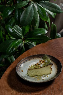 Fıstıklı kibrit peynirli kek ve seramik tabakta yeşil çay tozuyla kaplı hindistan cevizi cipsi. Masanın yanında bir bardak, bir şişe Japon şarabı ve bitkiler var..