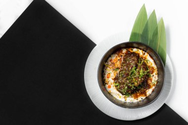 Kızarmış dana yanakları unagi soslu ve patates püresinin üstünde yeşil soğan var. Yiyecek koyu bir seramik kasede, bambu yapraklı hafif bir tabakta yatıyor. Tabaklar siyah beyaz bir arka planda duruyor..