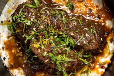 Kızarmış dana yanakları unagi soslu ve patates püresinin üstünde yeşil soğan var. Yiyecek koyu bir seramik kasede, bambu yapraklı hafif bir tabakta yatıyor. Tabaklar siyah beyaz bir arka planda duruyor..