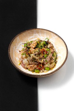 Deniz ürünleri, ton balığı, kalamar, somon, yeşil fasulye, pırasa ve mikroyeşil lahanalı pilav. Yiyecek, siyah ve beyaz arka planda kahverengi bir seramik tabakta yatıyor. Alçı figürünün yanında..