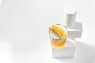 Şeffaf bir bardakta, üzerinde muz ve mango tozu olan şeffaf bir buz ve narenciye köpüğü ile mango ekşi kokteyli. Kokteyl, beyaz arka plandaki diğer figürler arasında alçı bir figürün üzerinde duruyor..