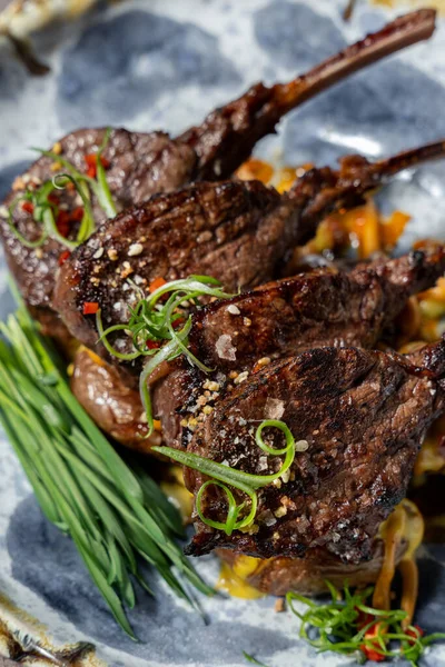 stock image Grilled rack of lamb with green onions and fried potatoes in mango sauce on a light ceramic plate with a blue pattern. The plate sits on a gray fabric chair.