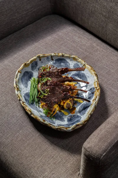 用青葱烤羊腿 用芒果酱煎土豆 放在蓝色图案的轻陶瓷盘上 盘子坐在一把灰色的布椅上 — 图库照片
